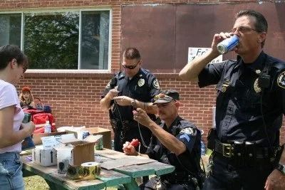 Police officers at an outdoor event, one drinks from a can, others work on equipment at a table.