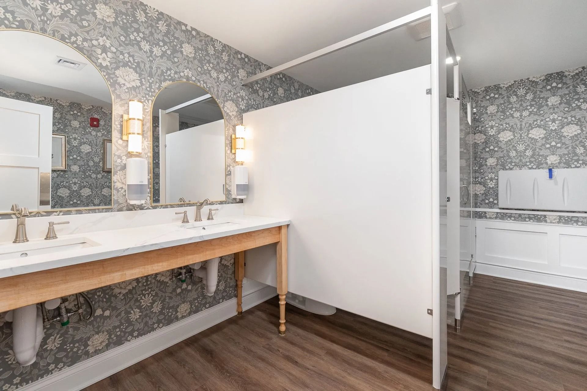 Public restroom with white stalls, double sinks, floral wallpaper, and wood-look flooring.