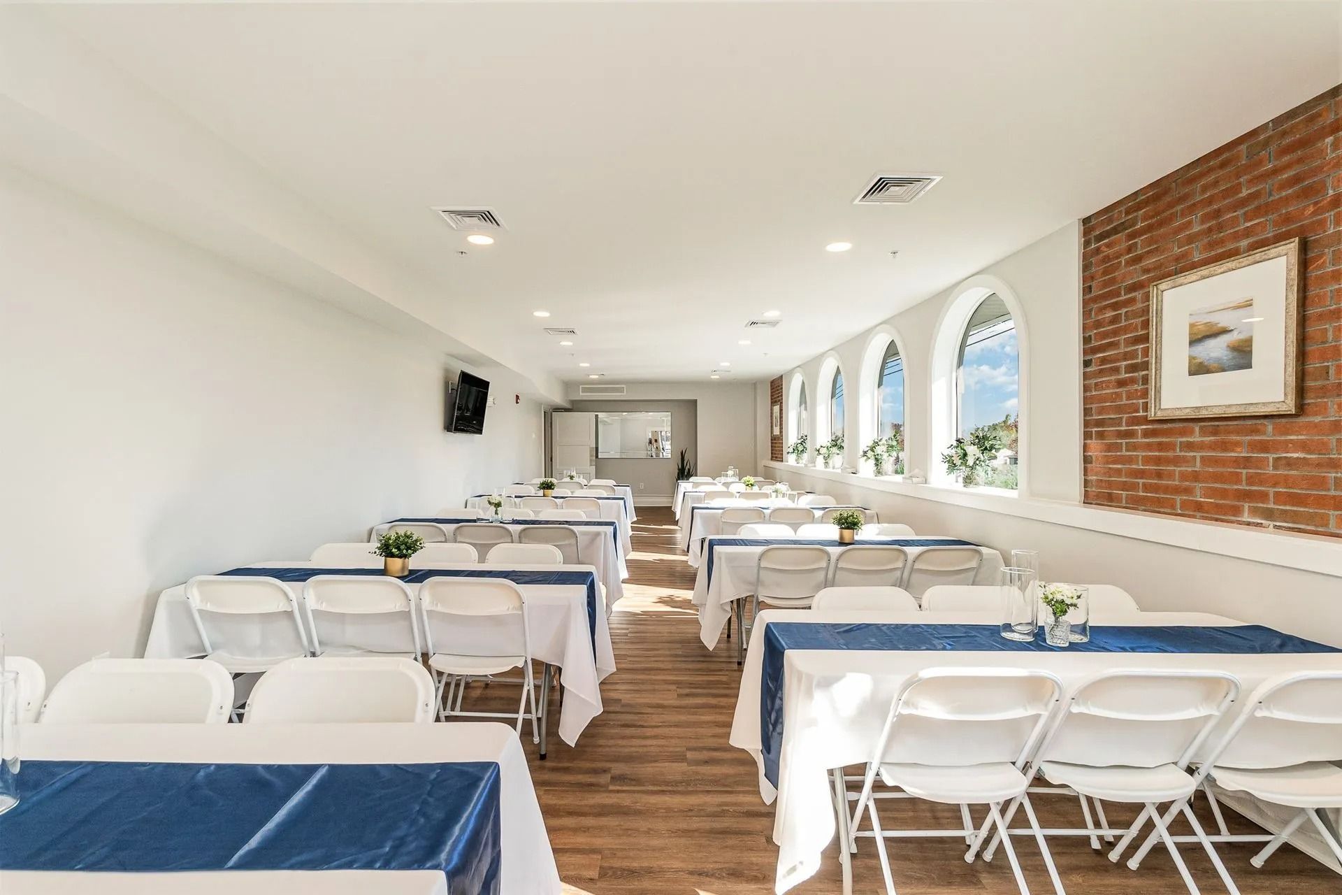 Interior of an event space with white tables, chairs, and a brick wall. Sunlight streams through arched windows.