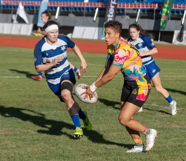 Rugby player in colorful jersey runs with the ball as two blue-and-white opponents chase on the field