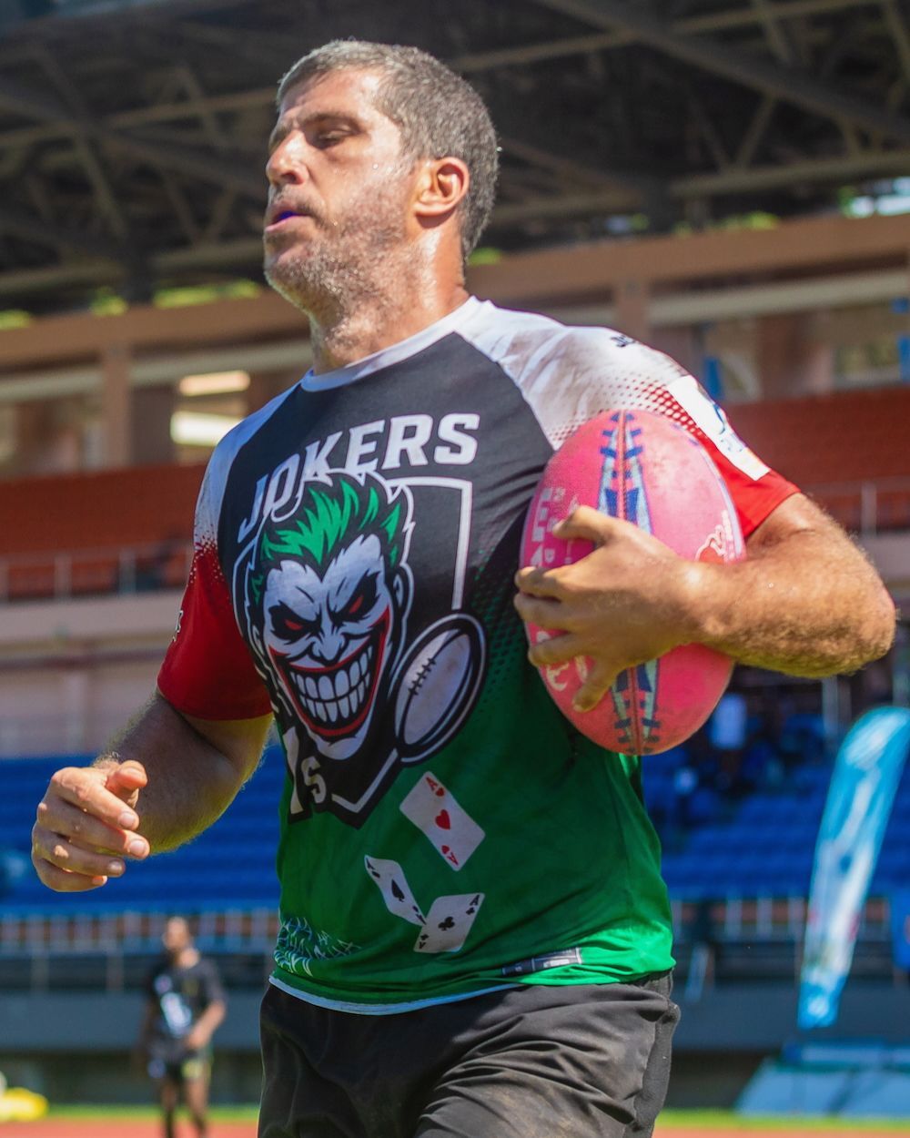 A runner in a green and black JOKERS shirt carries a red rugby ball on a stadium field.