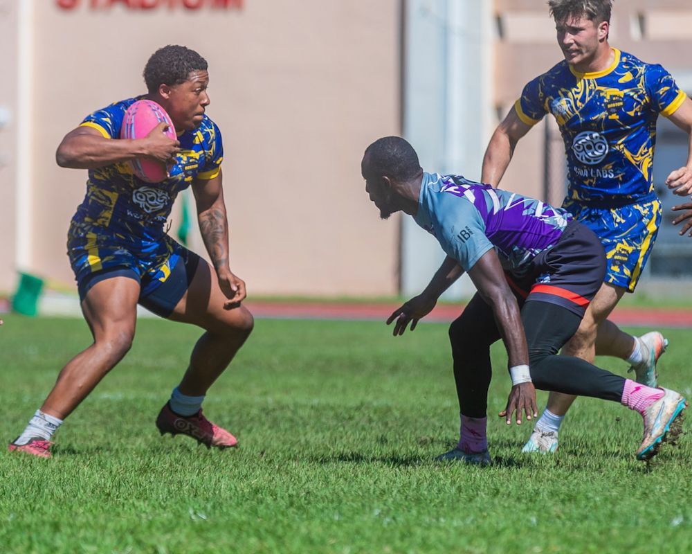 Rugby players in blue and purple jerseys crouch and brace for contact on a grassy field.