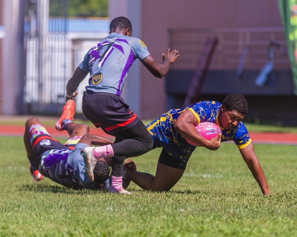 Rugby player in blue and yellow dives forward as two opponents tackle him on a grassy field