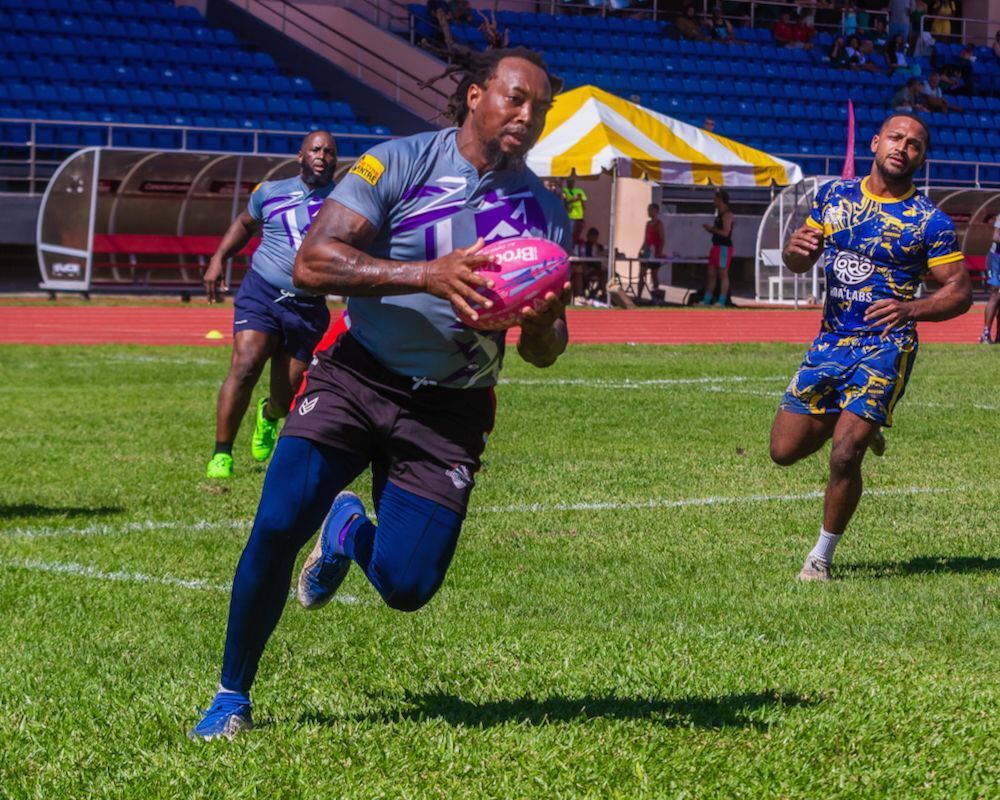 Rugby player in gray and blue runs with the ball on a sunny field, with opponents chasing behind.