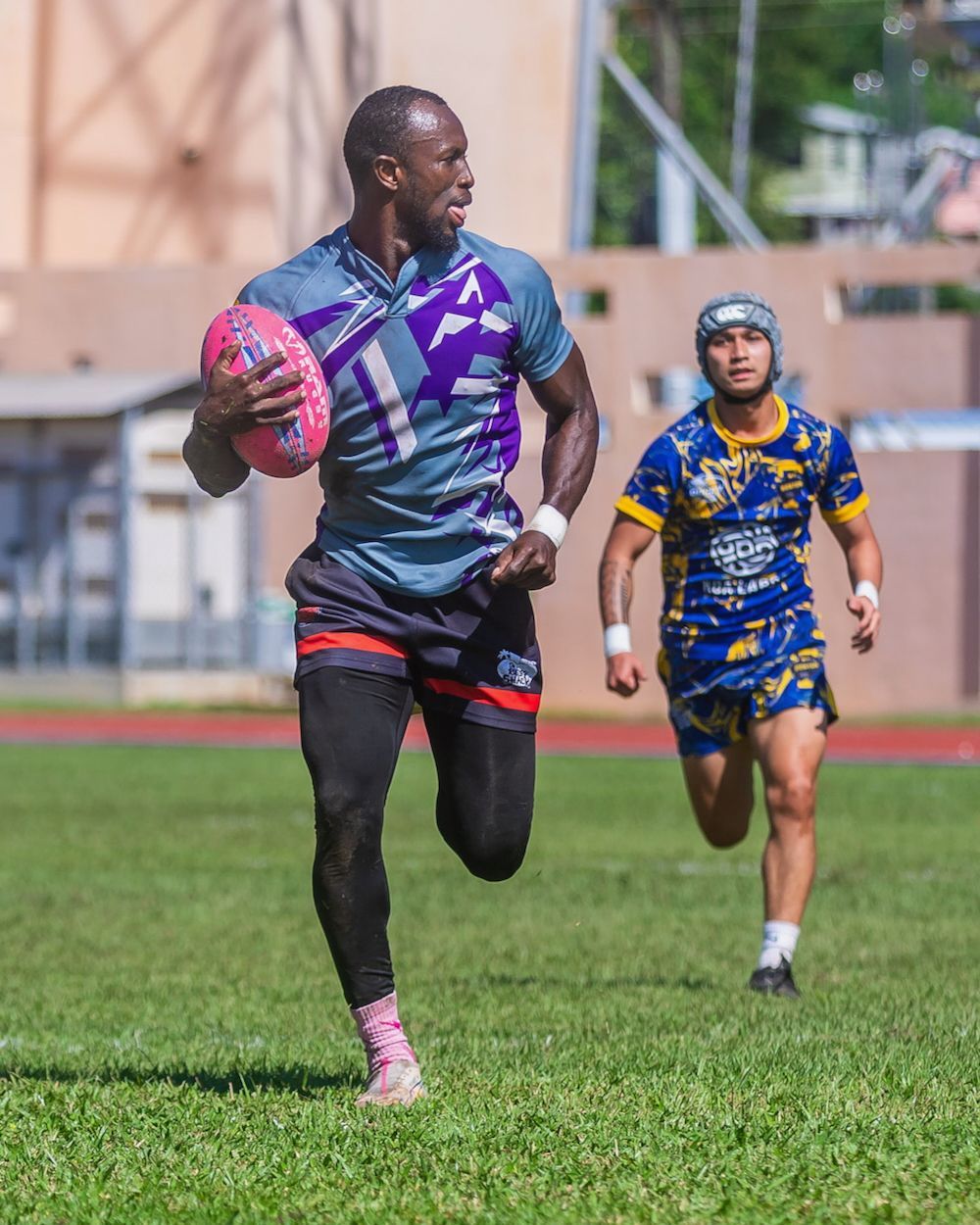 Rugby player in purple and black sprints with the ball while an opponent in blue chases on a grassy field.