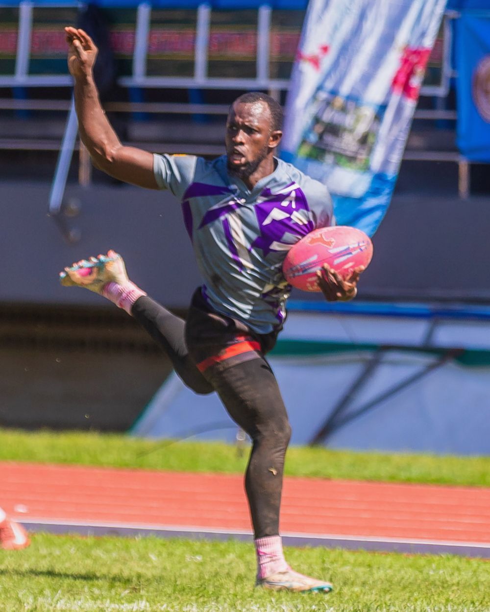 Athlete in black and purple mid-kick on a rugby field, holding a pink ball, with one arm raised
