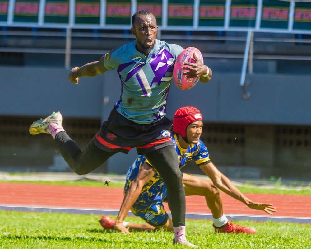 Rugby player in purple leaps past a defender on a field, carrying the ball during a match.