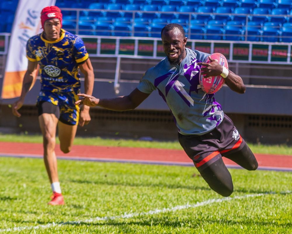 Two athletes sprint on a track, one in purple and black leaning forward with a ball, another chasing behind.