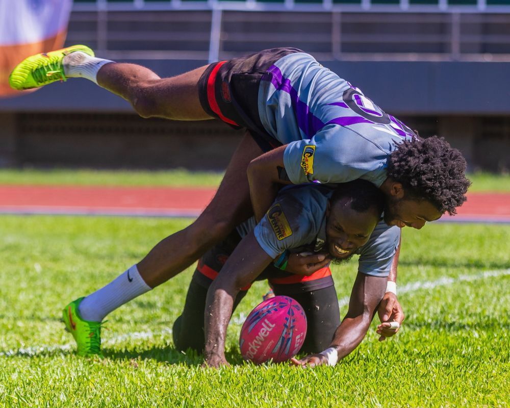 Two rugby players tackle over a pink ball on a grass field, one airborne in a stadium.