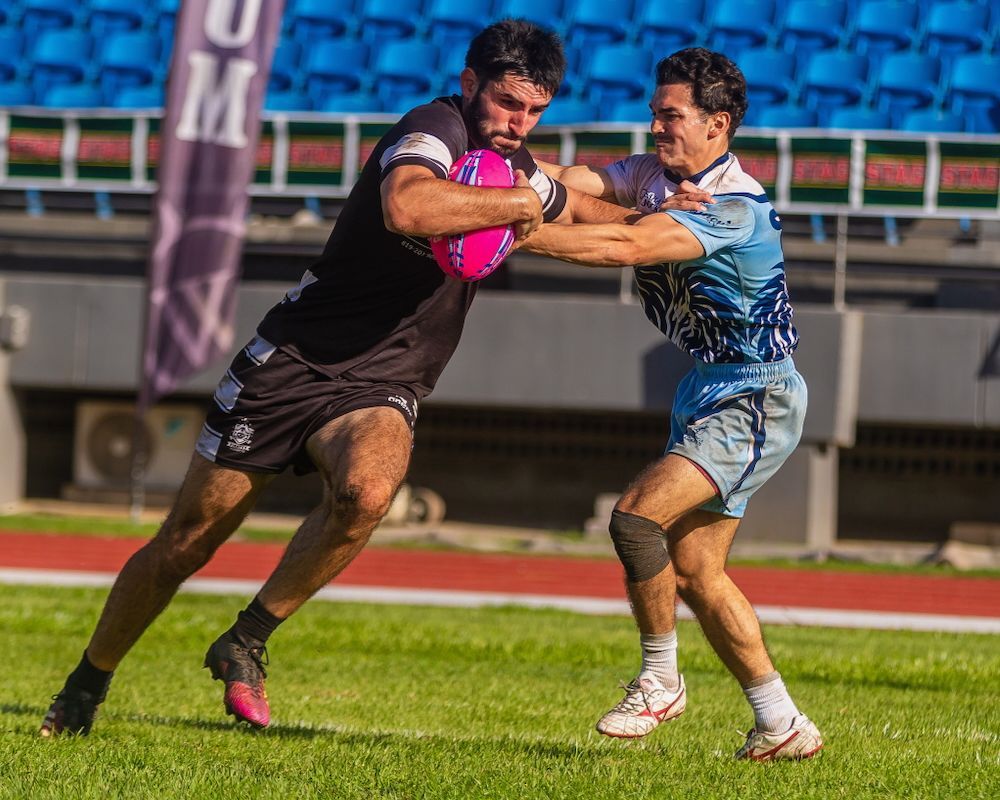 Two rugby players in black and blue jerseys collide during a tackle on a grassy field.