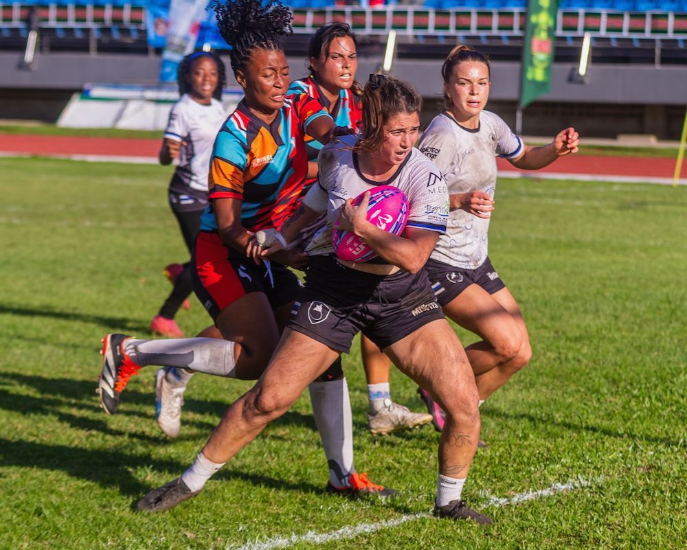 Rugby players sprint in a close chase on a grassy field, with one carrying a pink ball.