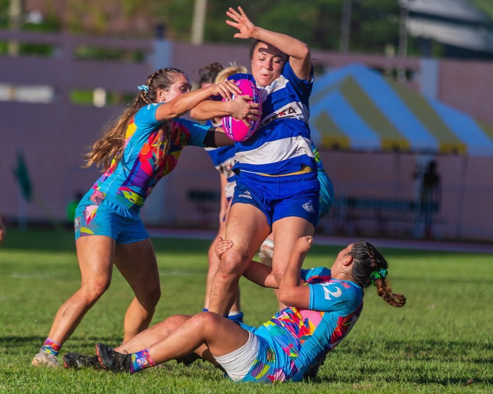 Rugby players in blue and pink jerseys tackle on a grass field during a match.
