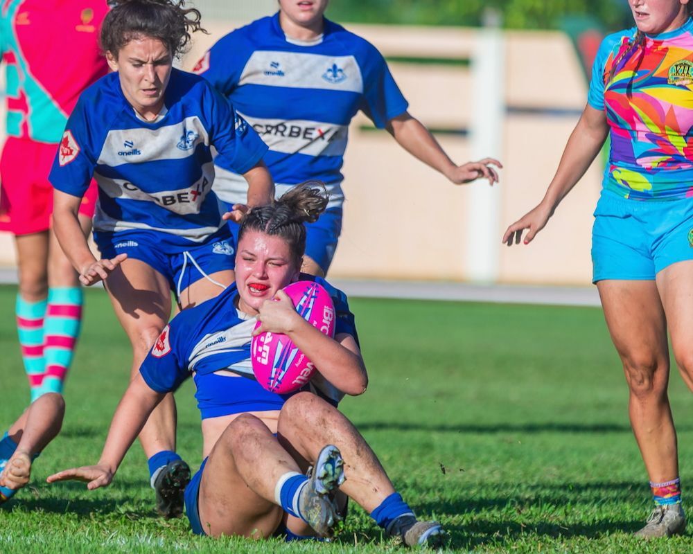 Rugby player in blue and pink slides on grass, clutching the ball as opponents chase behind.