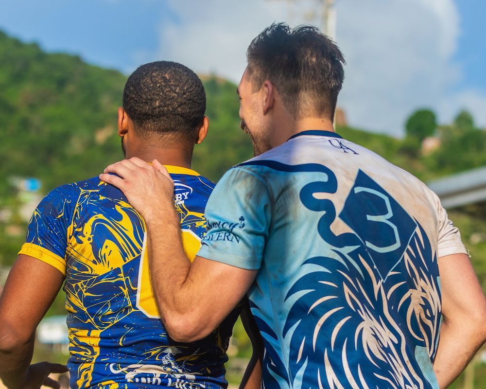 Two athletes in blue jerseys stand side by side outdoors, one with an arm around the other’s shoulder.