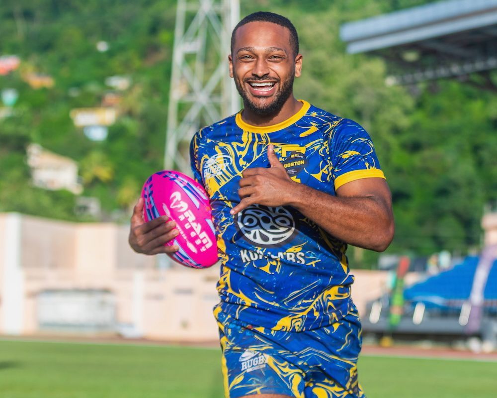 Smiling athlete in blue-and-yellow uniform holding a pink rugby ball on a field