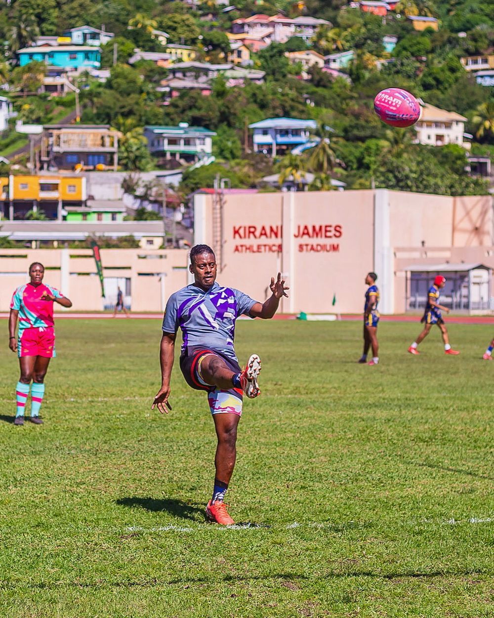 Rugby player in purple kicks the ball on a grassy field, with teammates and hillside houses in the background.