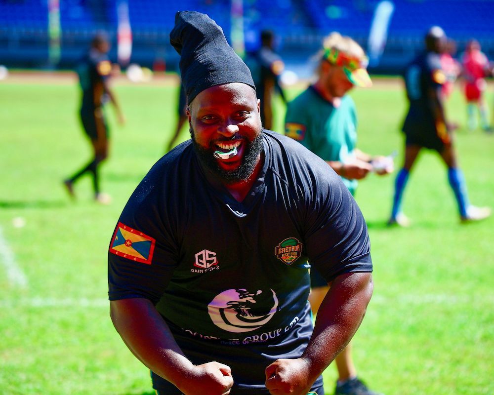 Smiling athlete in black jersey and beanie kneels on a grassy sports field during daylight