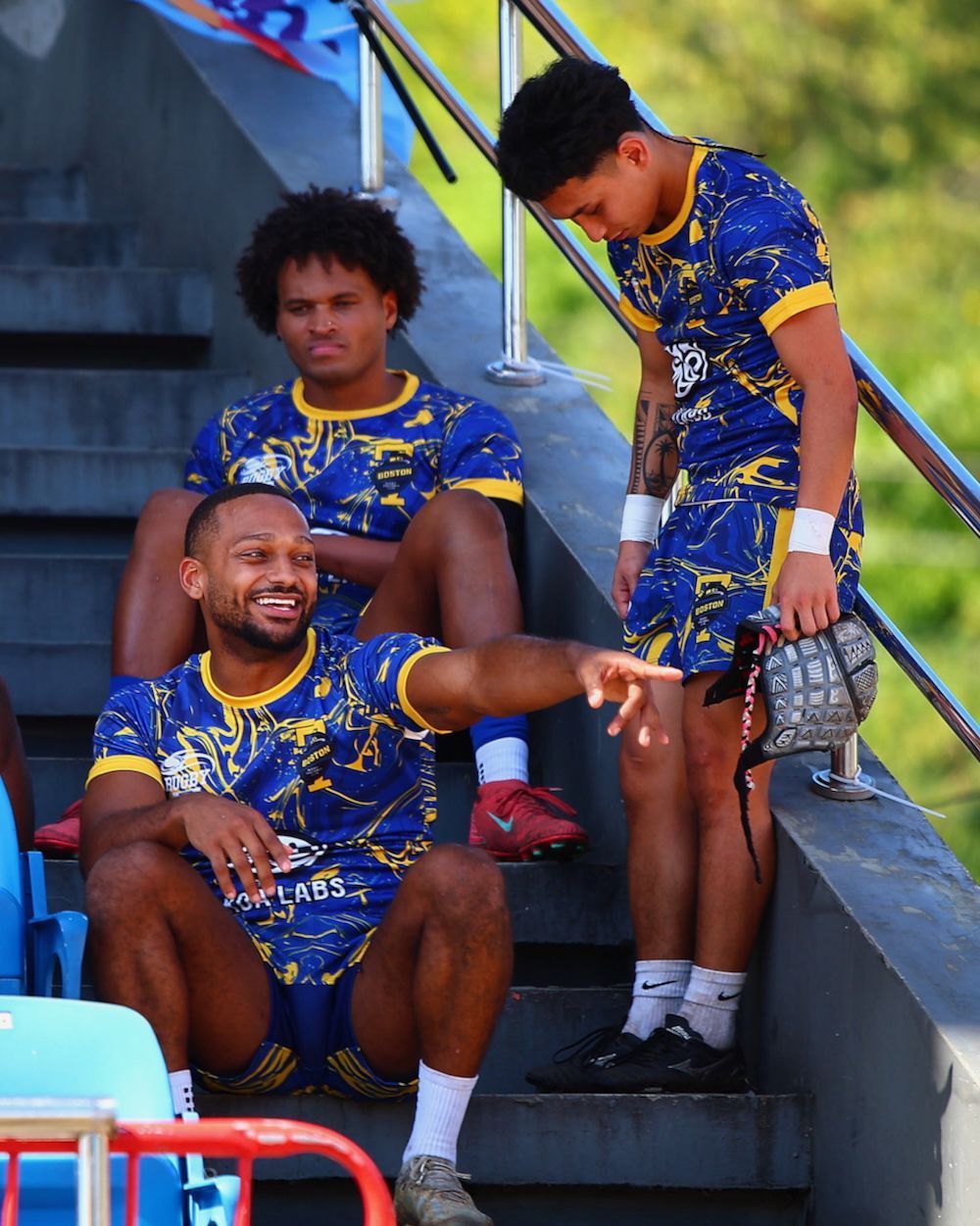 Three athletes in blue-and-gold uniforms sit on stadium steps, one smiling and pointing.