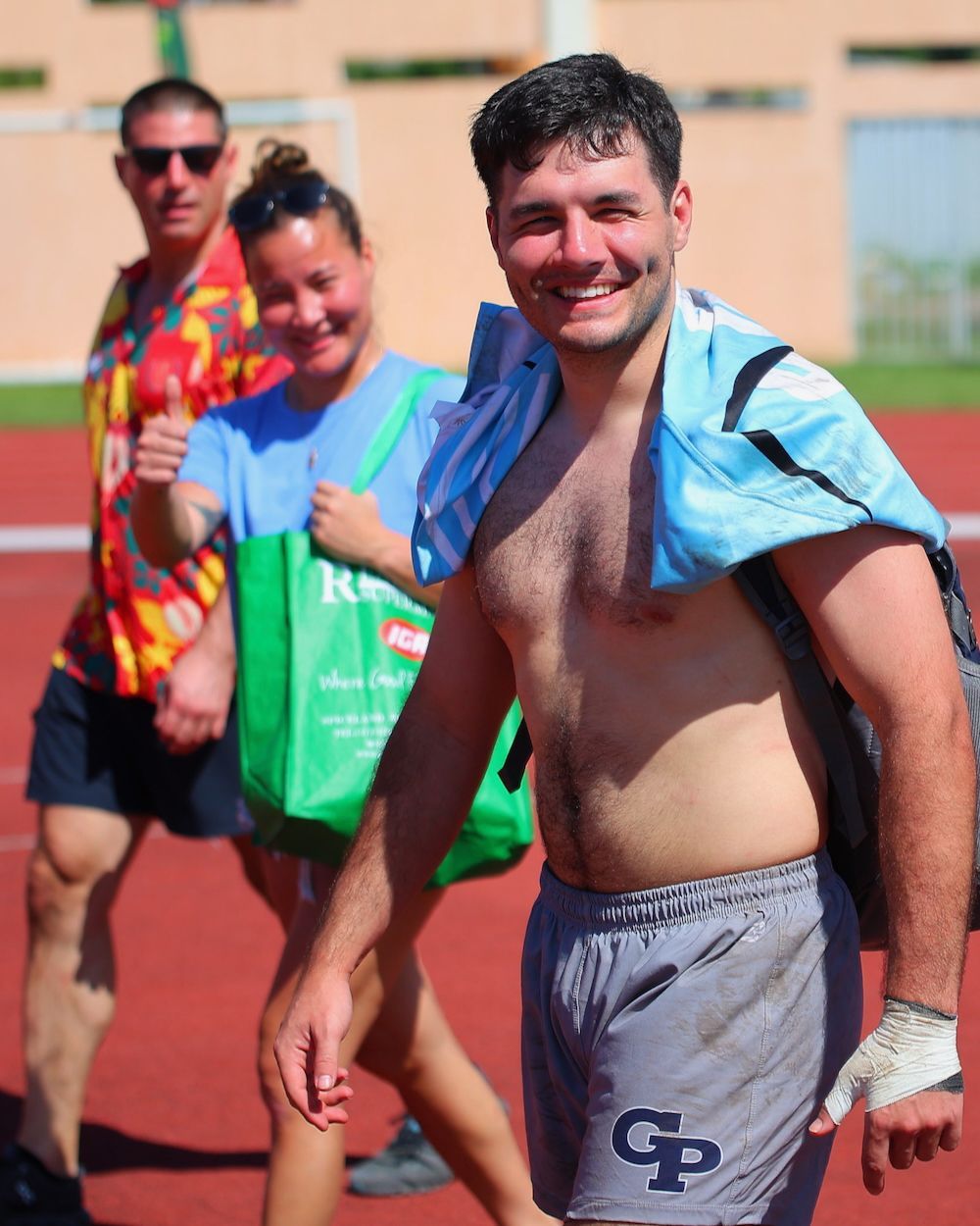 Smiling man in swim trunks and a blue towel walks outdoors beside two others near a track