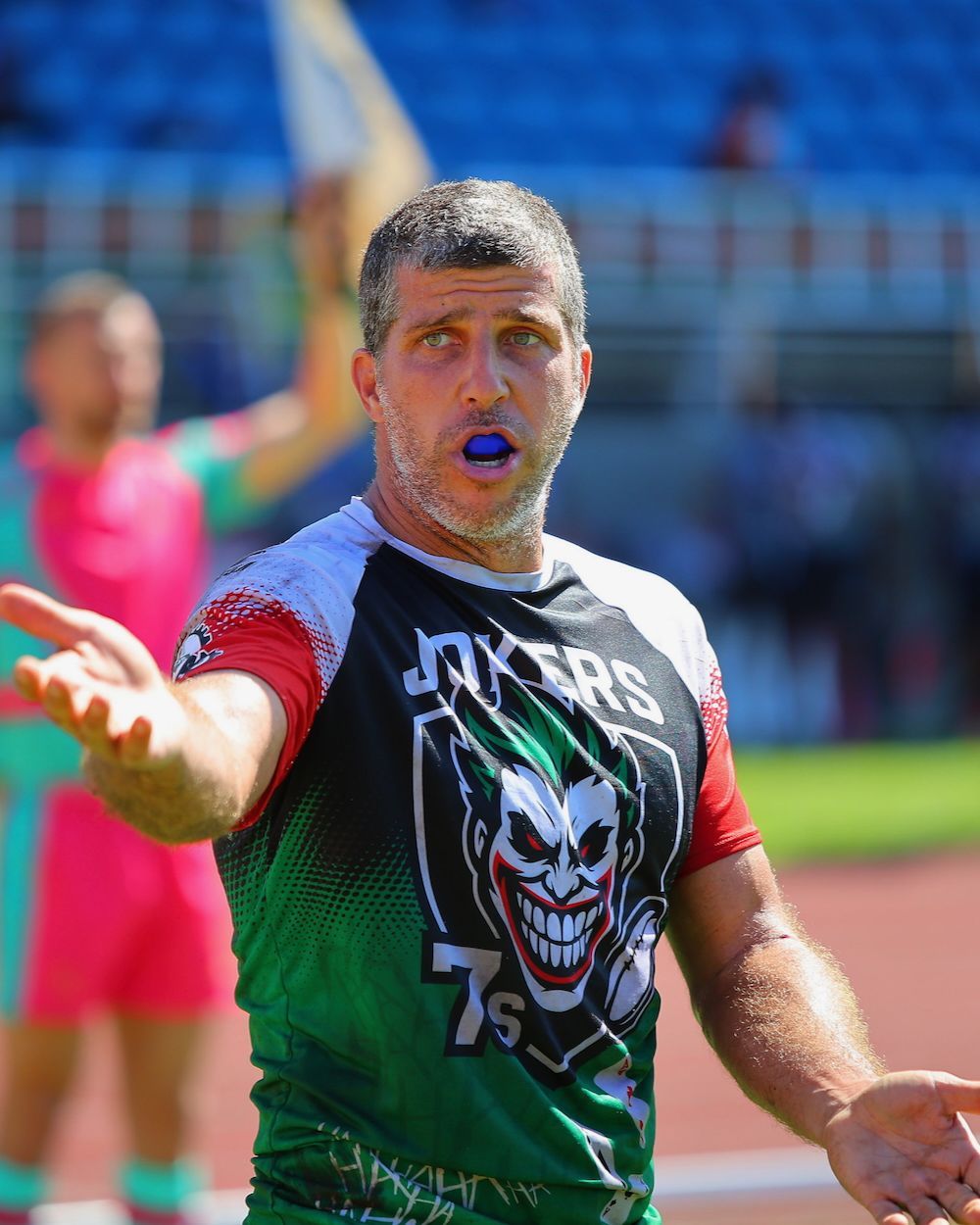 Athlete in a green and black jersey with a Joker graphic, gesturing on a sports field, blurred stadium behind him