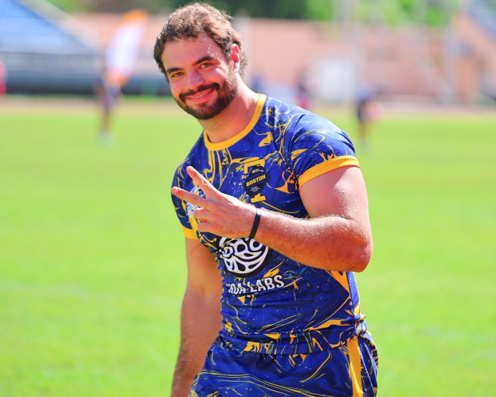 Smiling athlete in blue and yellow sports uniform making a peace sign on a grassy field