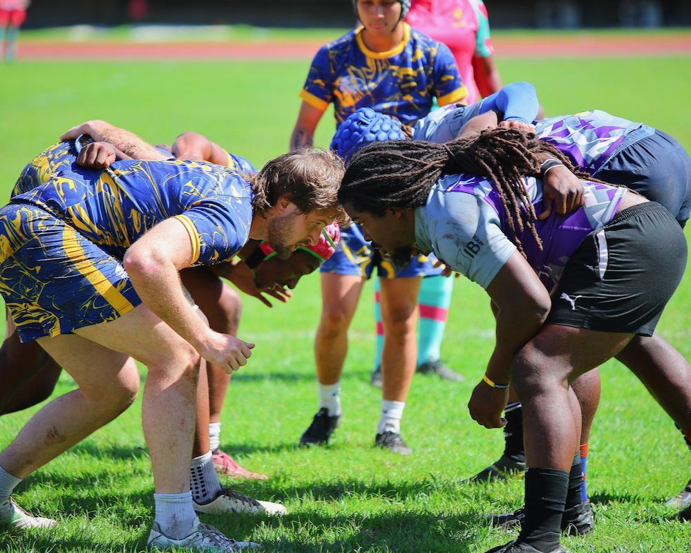 Two rugby players crouch face to face on a grassy field during a scrum, with a referee standing behind them.