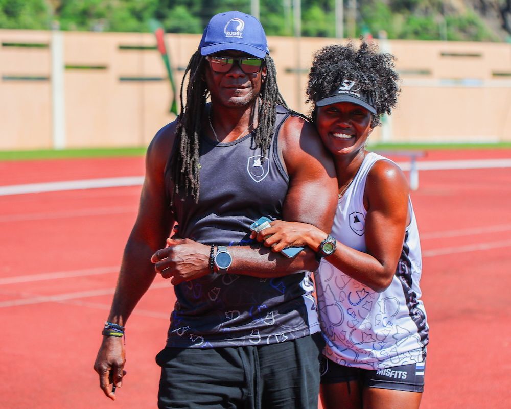 Two athletes smiling and posing on a red track, one in a blue cap and one in a white tank top.