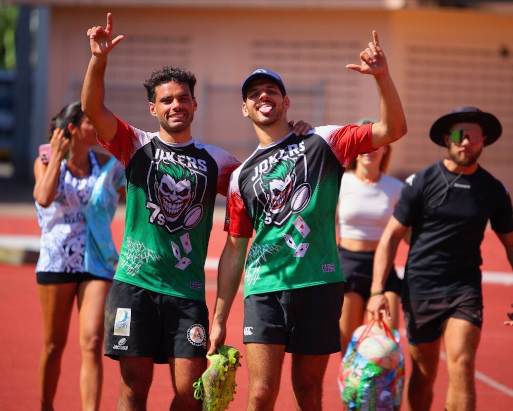 Two runners in matching green jerseys pose with raised hands on a track, smiling in bright sunlight