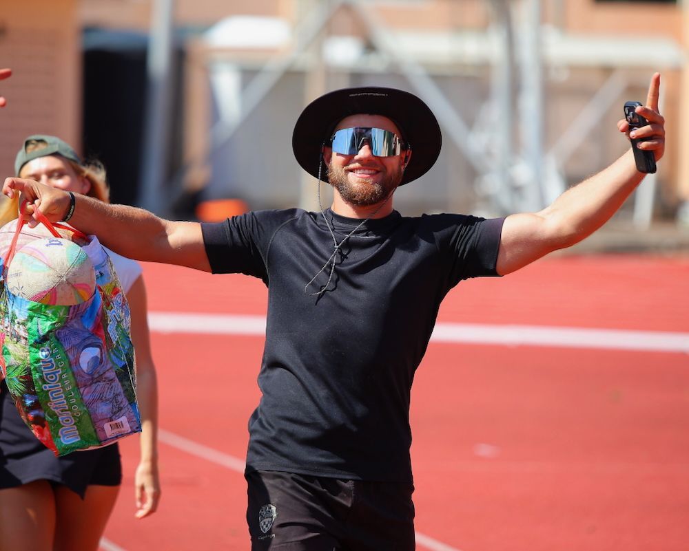 Person in black shirt and hat with mirrored sunglasses, arms outstretched on a red track in bright sunlight