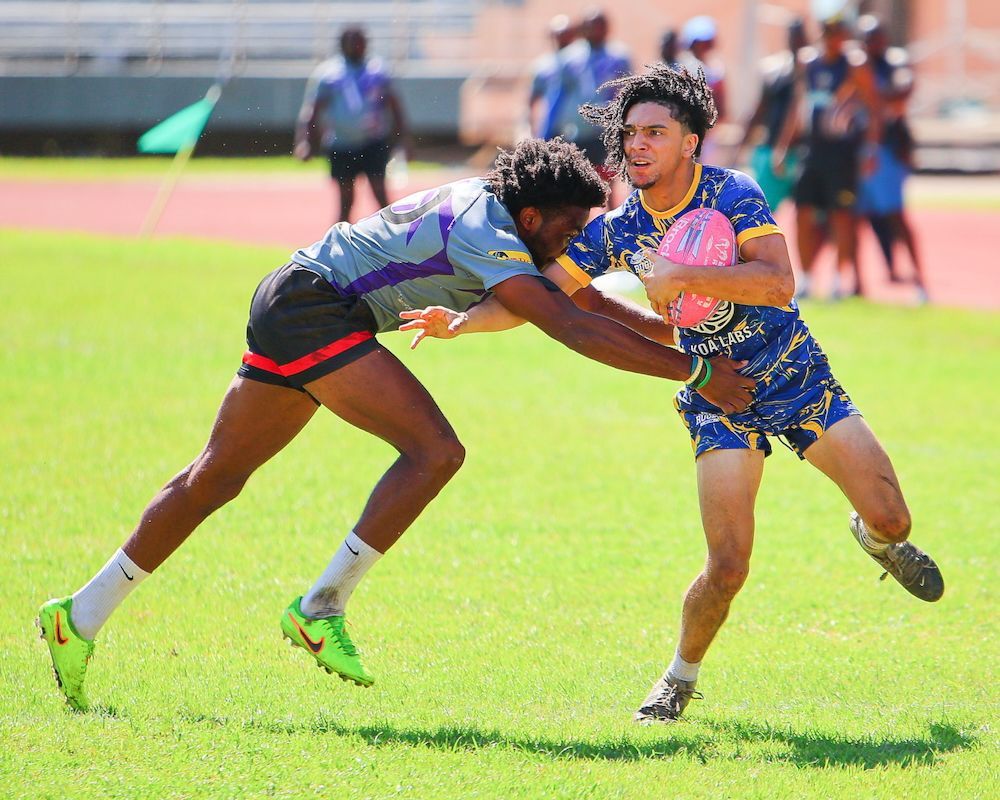 Two athletes in colorful uniforms grapple on a grassy track field during a game.