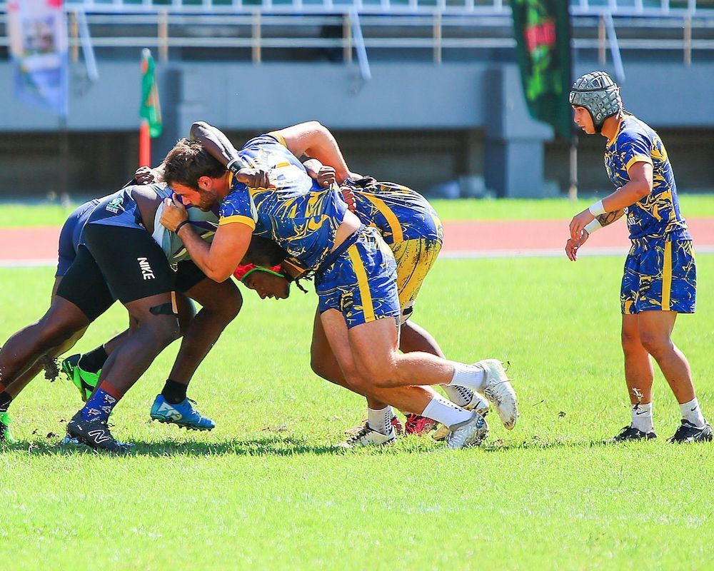 Two rugby players in blue and yellow tackle an opponent on a grassy field.