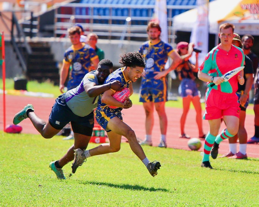 Rugby players sprint and tackle on a bright grass field with spectators in the background
