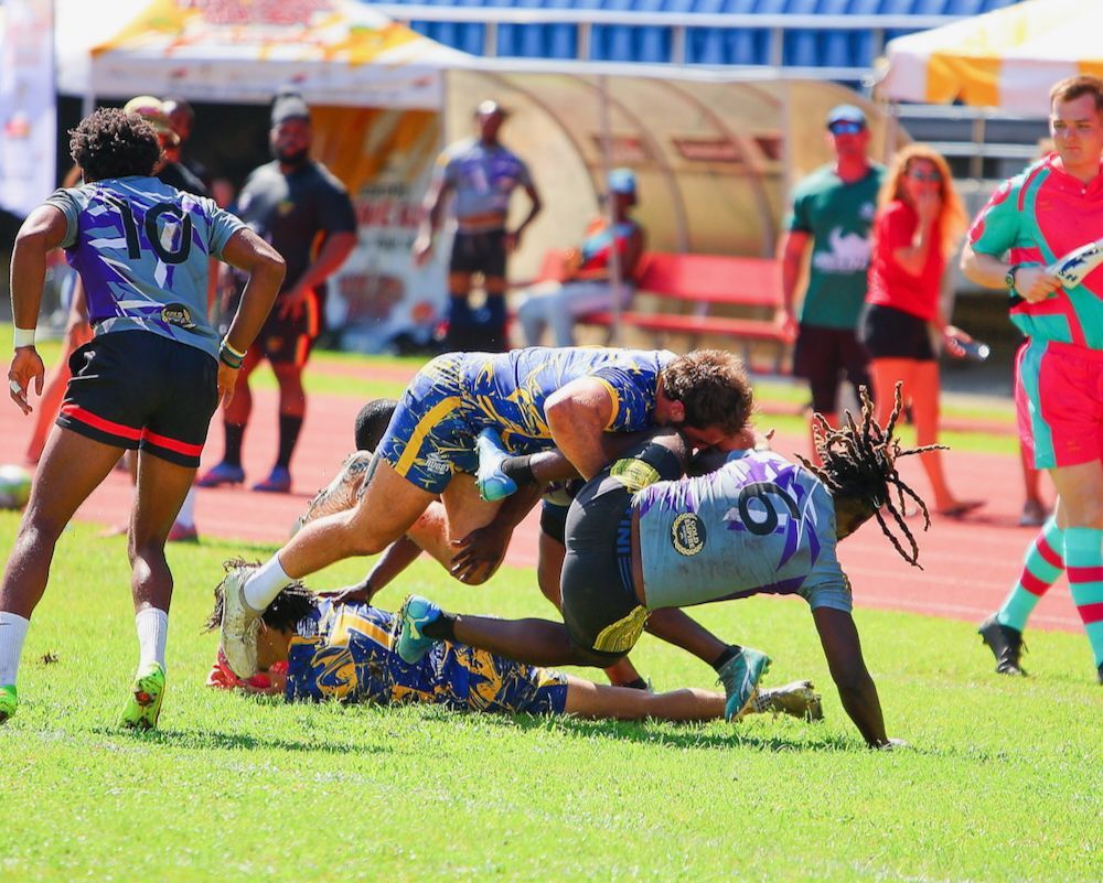 Rugby players tackling on a sunny field, with one player on the ground and others piled over the ball.