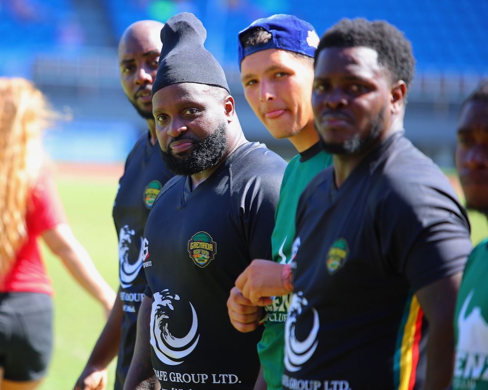 Four athletes in black training shirts and caps stand on a sunny sports field.