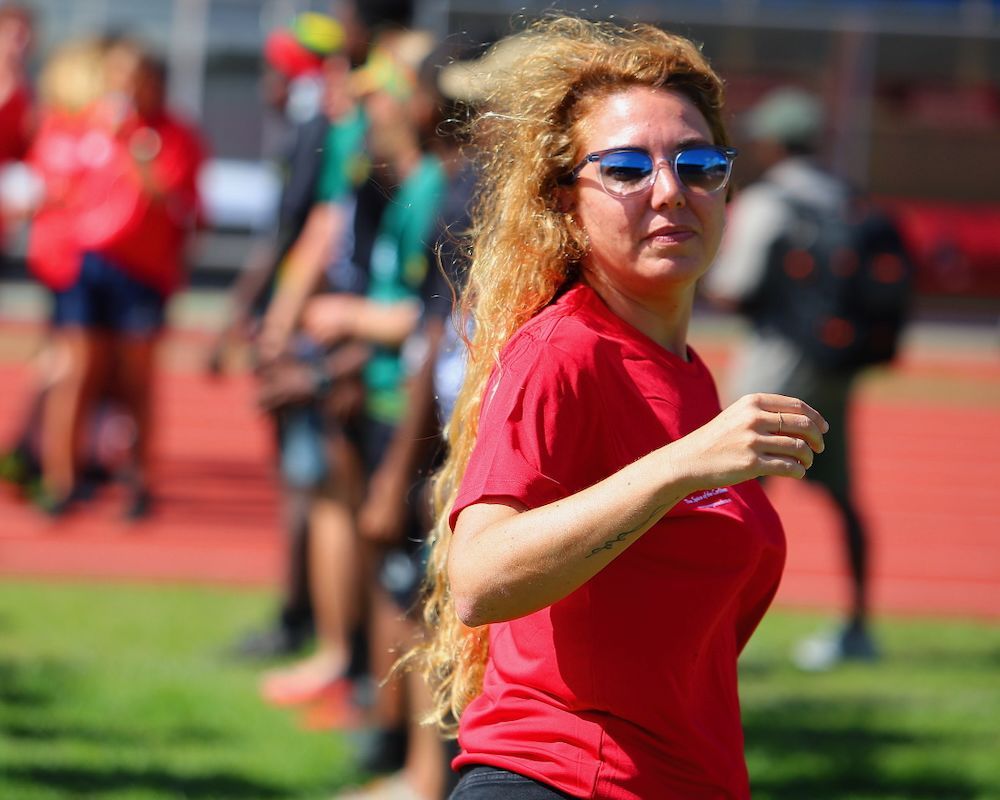 Woman in red shirt and blue sunglasses standing on a track field, with blurred people in the background