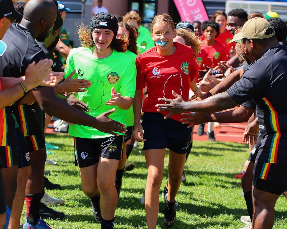 Runners in bright shirts run through a cheering crowd, hands outstretched in a sunny outdoor race event.