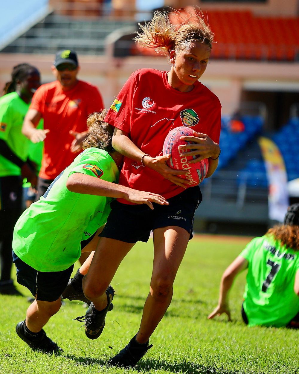 Rugby player in red runs with the ball as a player in green tackles on a grassy field.