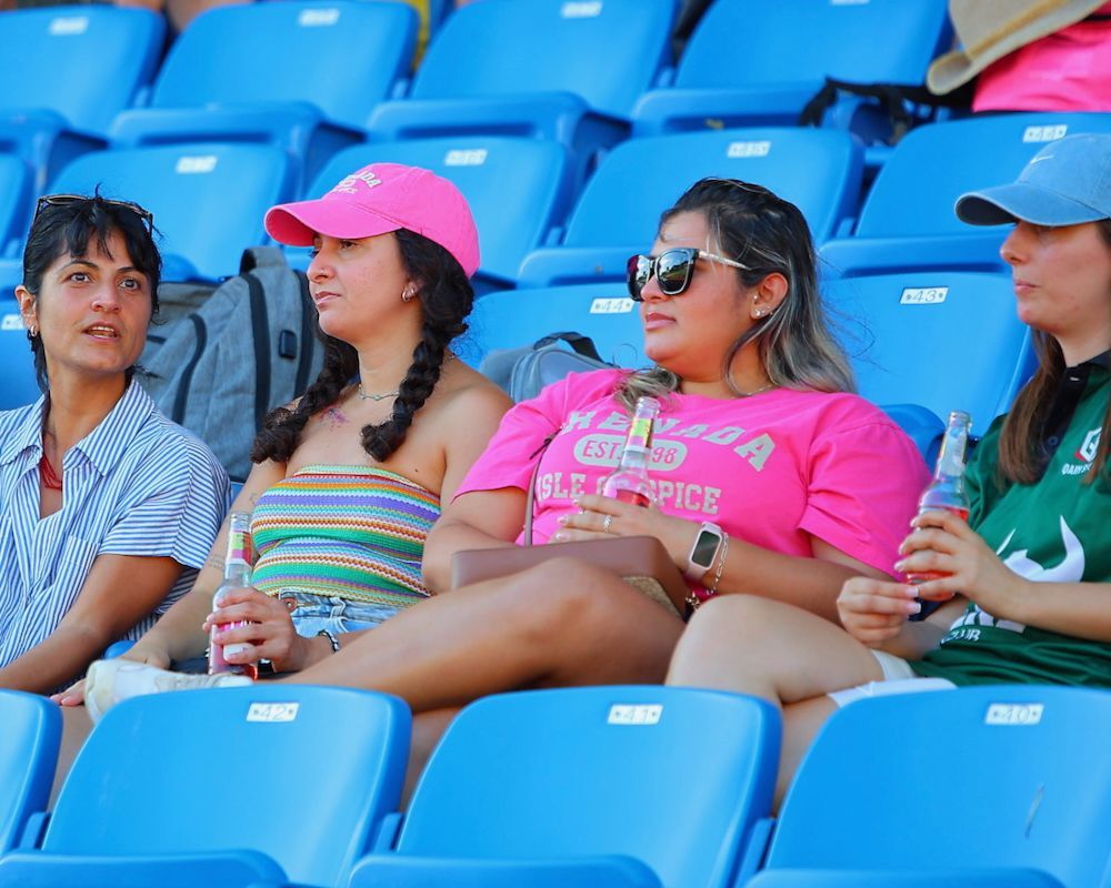 Four people sitting in blue stadium seats, chatting and wearing bright hats and sunglasses.