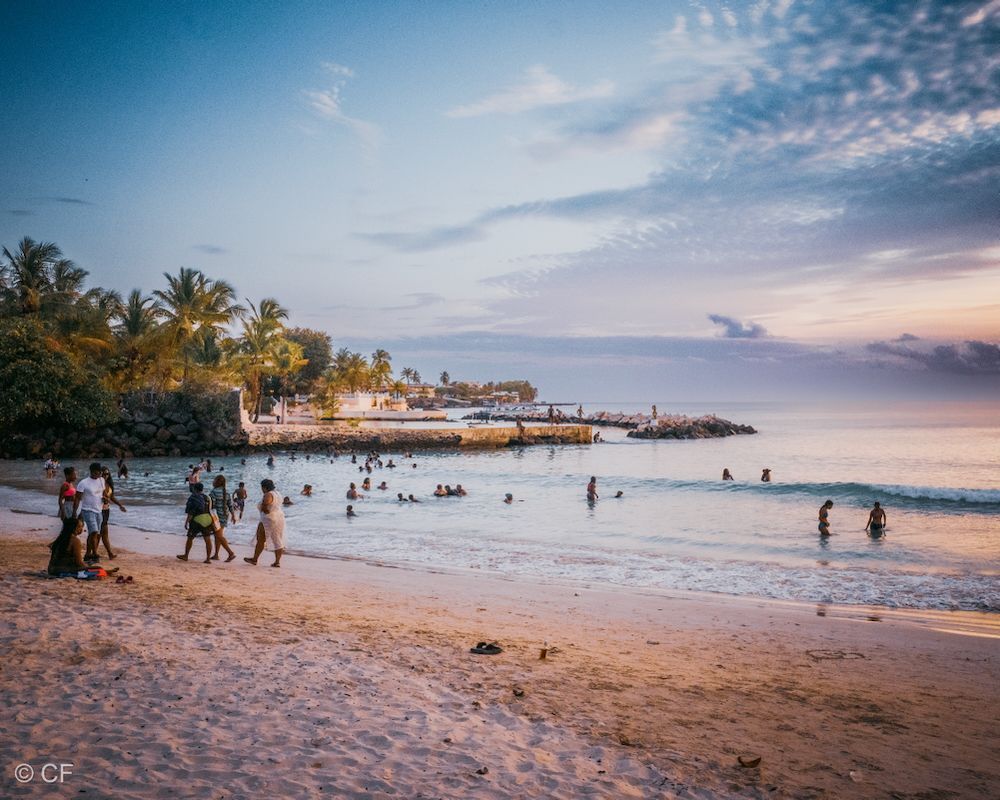 Beach at sunset with people wading in calm water, palm trees, and a pier in the distance