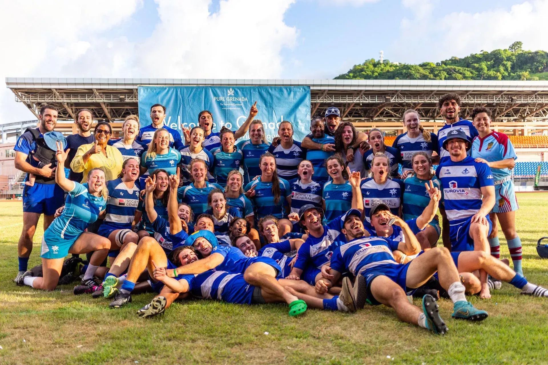 Large rugby team posing on a field, wearing blue and yellow uniforms in front of a stadium banner.