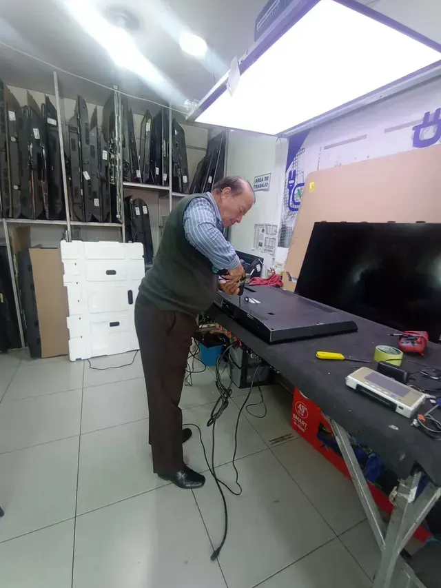 Un hombre trabaja en un banco de reparación desordenado en un taller, con herramientas y equipos en las paredes.
