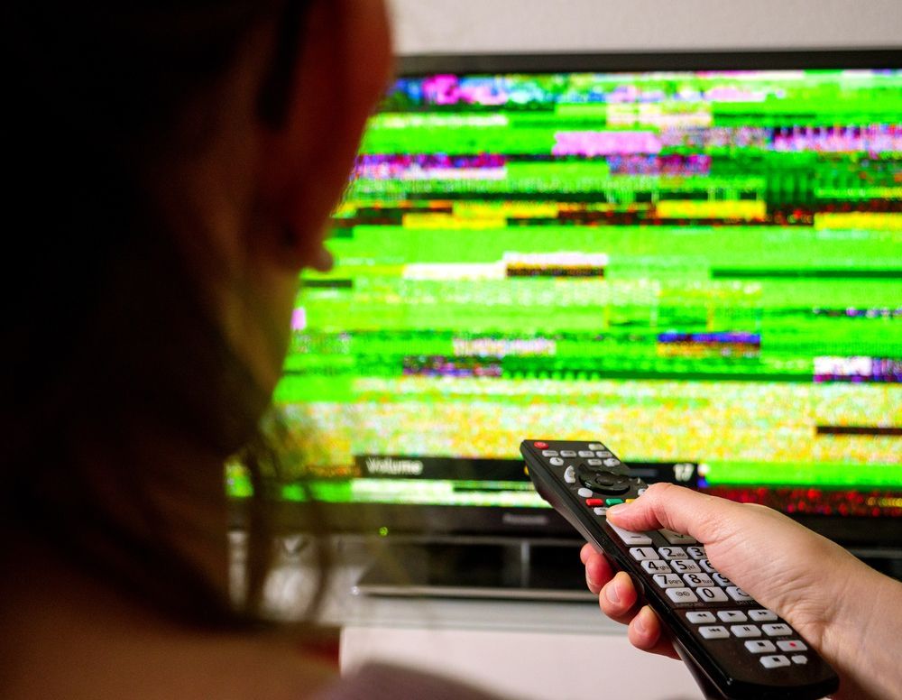 Una mano sostiene un control remoto apuntando hacia un televisor con una pantalla verde y con fallos.