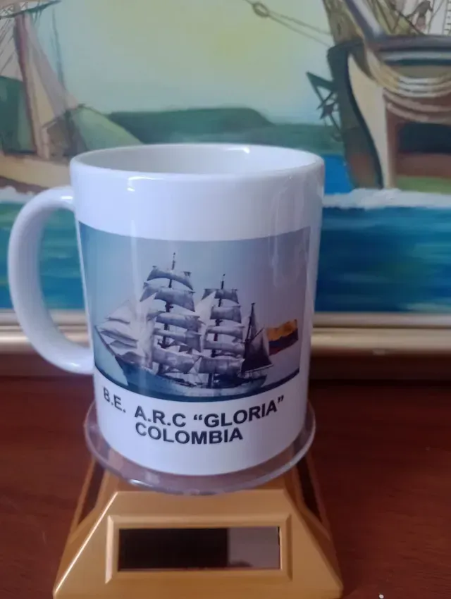Taza blanca con un velero y las palabras “ARC GLORIA COLOMBIA” sobre una base de madera.
