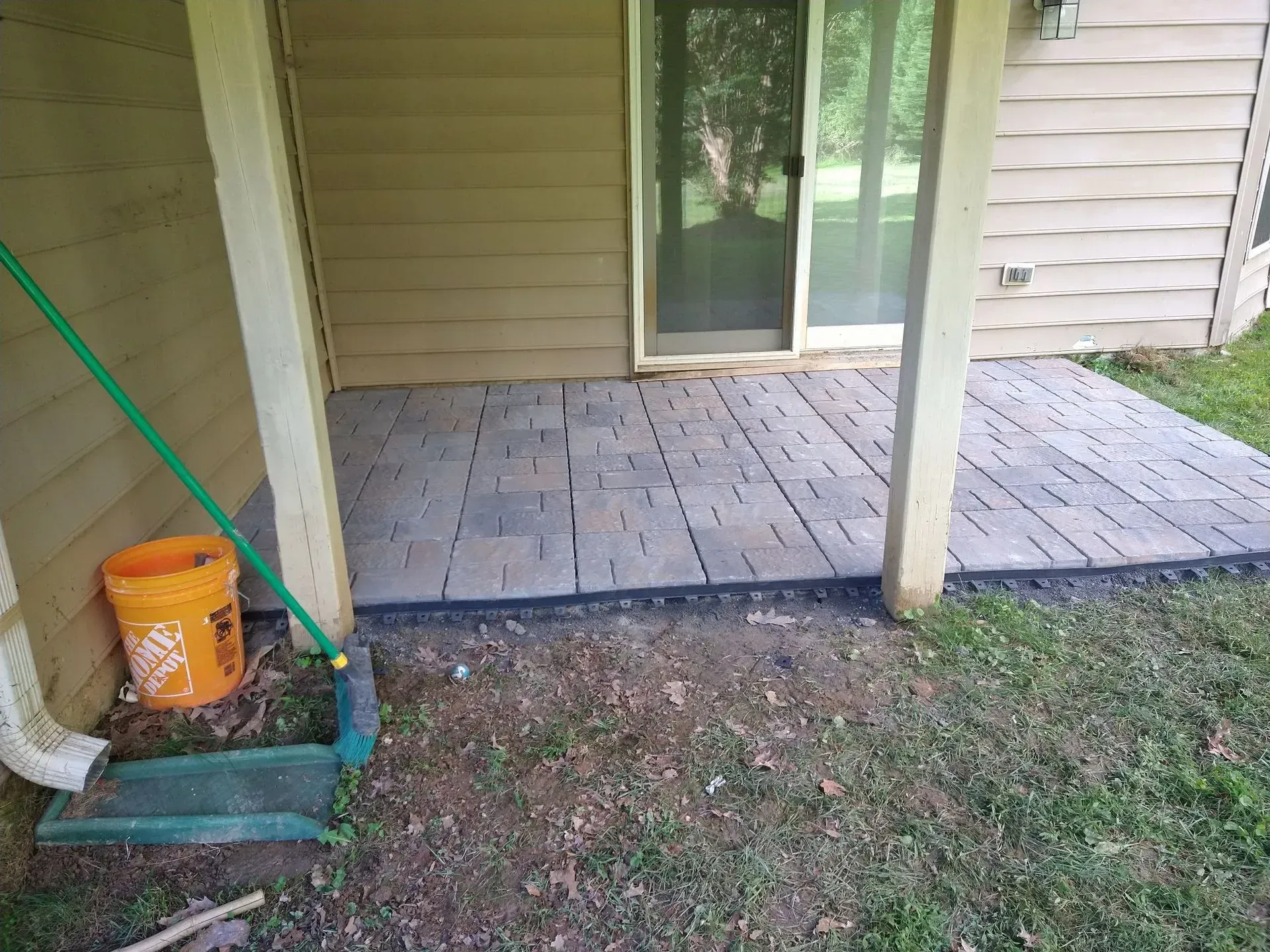 Back patio with square pavers, beige siding, a sliding glass door, and cleaning tools by the left post.