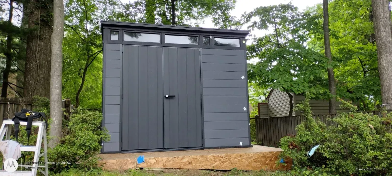 Gray backyard shed with double doors, surrounded by trees and greenery