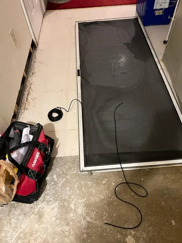 Black screen or panel leaning against a wall on a cluttered floor with cables and a red tool bag nearby