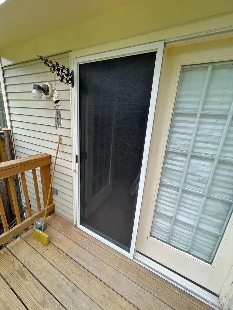 Outdoor porch with a black screen door beside white shutters and beige siding, wooden deck railing on left.