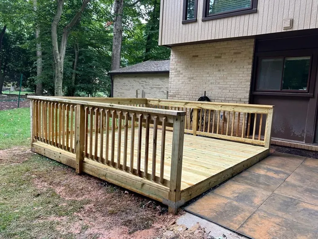 Wooden backyard deck with railings beside a house and patio, surrounded by trees.
