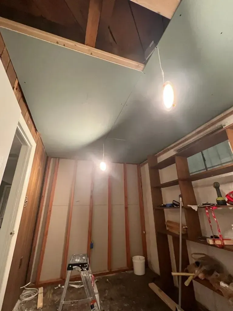 Basement room under construction with exposed insulation, ladder, shelves, and hanging lights.
