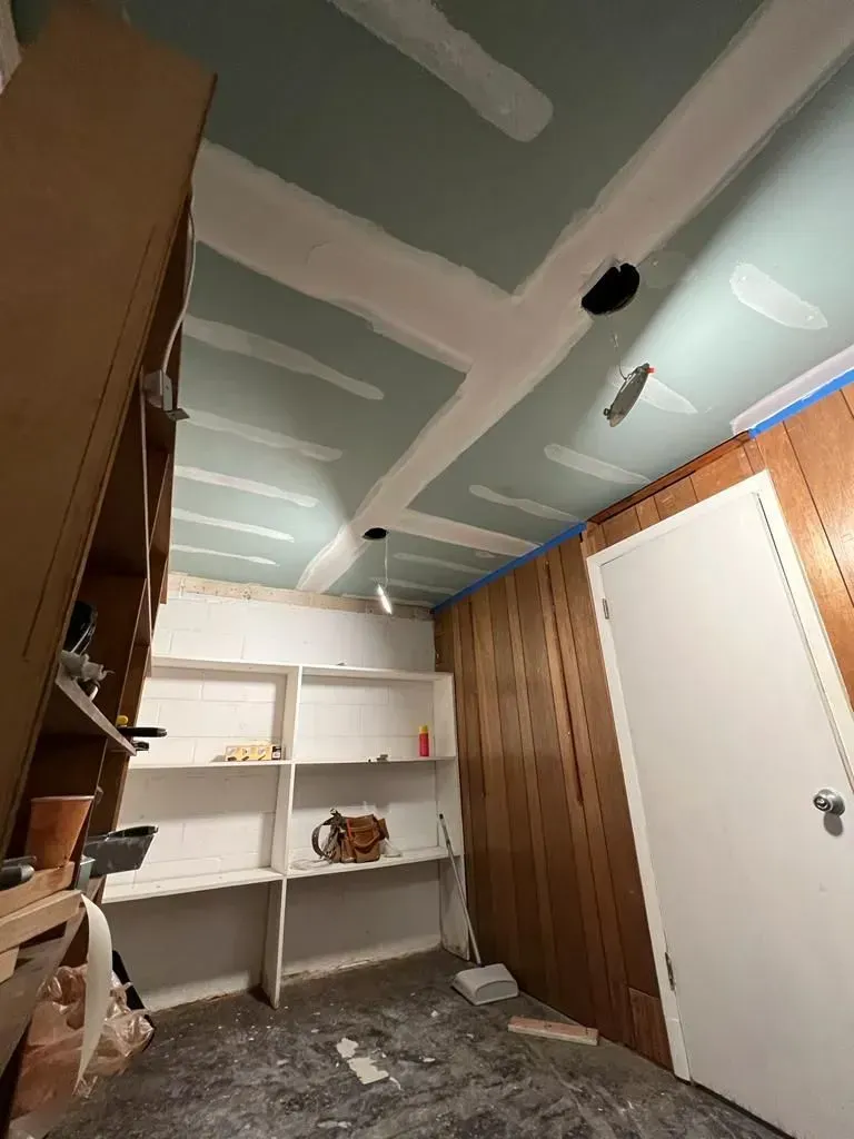 Small unfinished room with white shelves, wood-paneled walls, a white door, and exposed drywall on the ceiling.